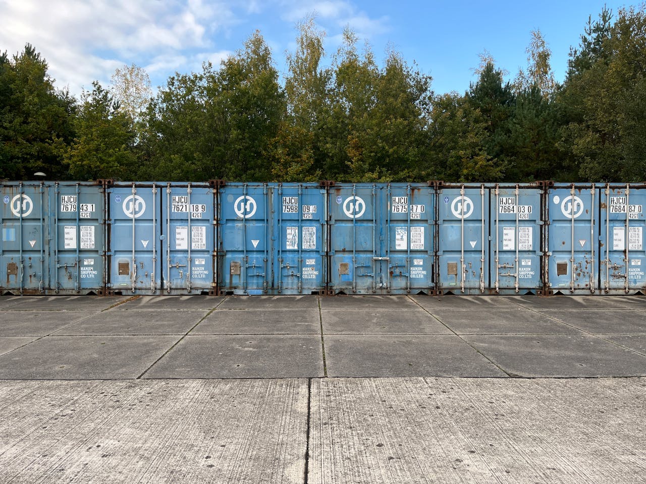 Home Blue shipping containers lined up outdoors against a forest background.