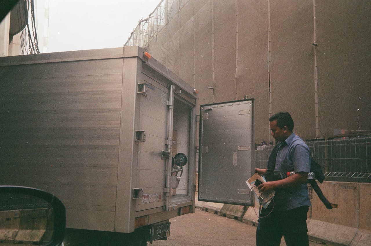 A logistics worker in a blue shirt checking a cargo truck on a city street. Industrial background suggests transportation and delivery.
